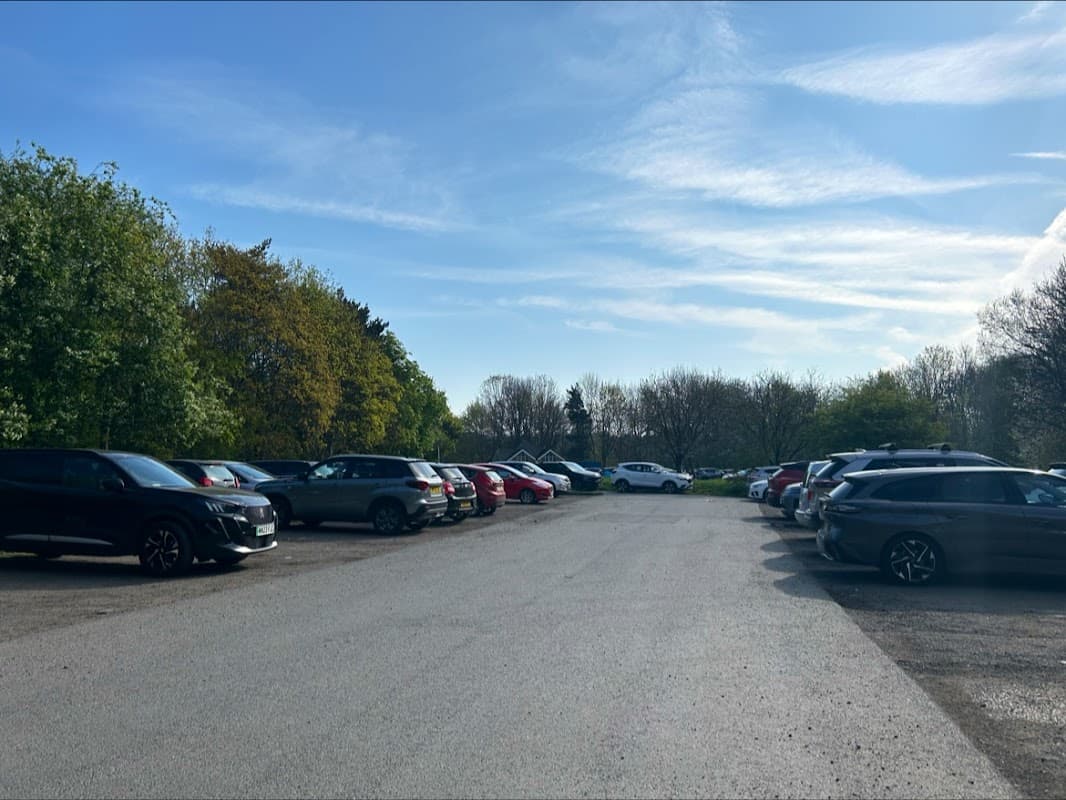 A spacious car park with various parked cars, surrounded by trees and a clear blue sky in Liversedge, Yorkshire.