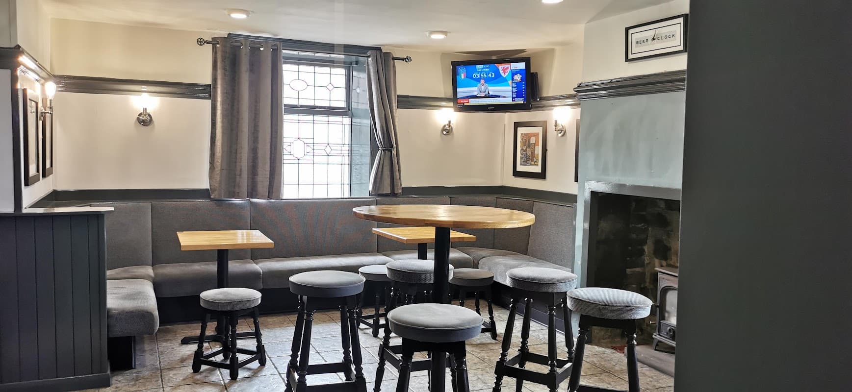 Cozy bar interior with wooden tables, high stools, a TV, and a fireplace, featuring gray walls and curtains.