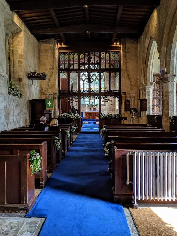 Interior of St Mary's Church with wooden pews, blue carpet, and stained glass windows. Floral decorations line the aisle.