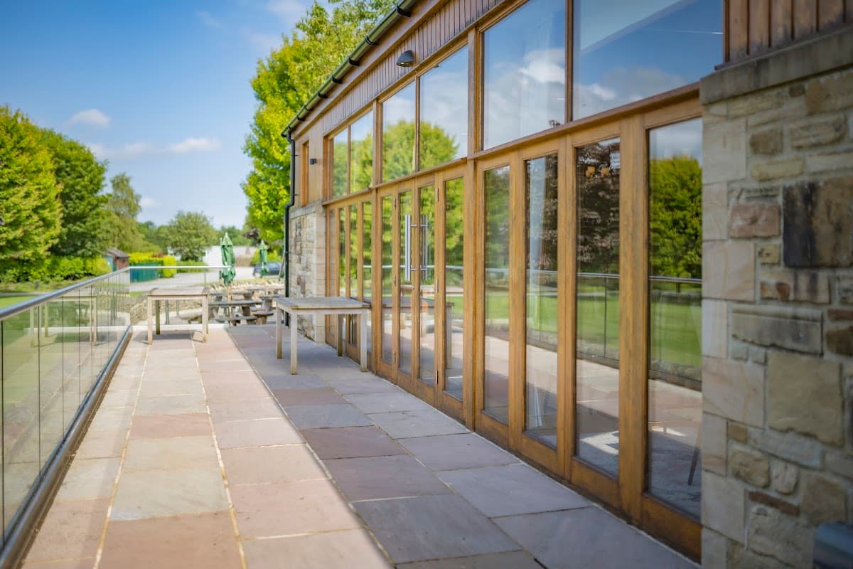 Wooden-framed glass doors open to a patio area with stone tiles, surrounded by lush green trees and a clear blue sky.