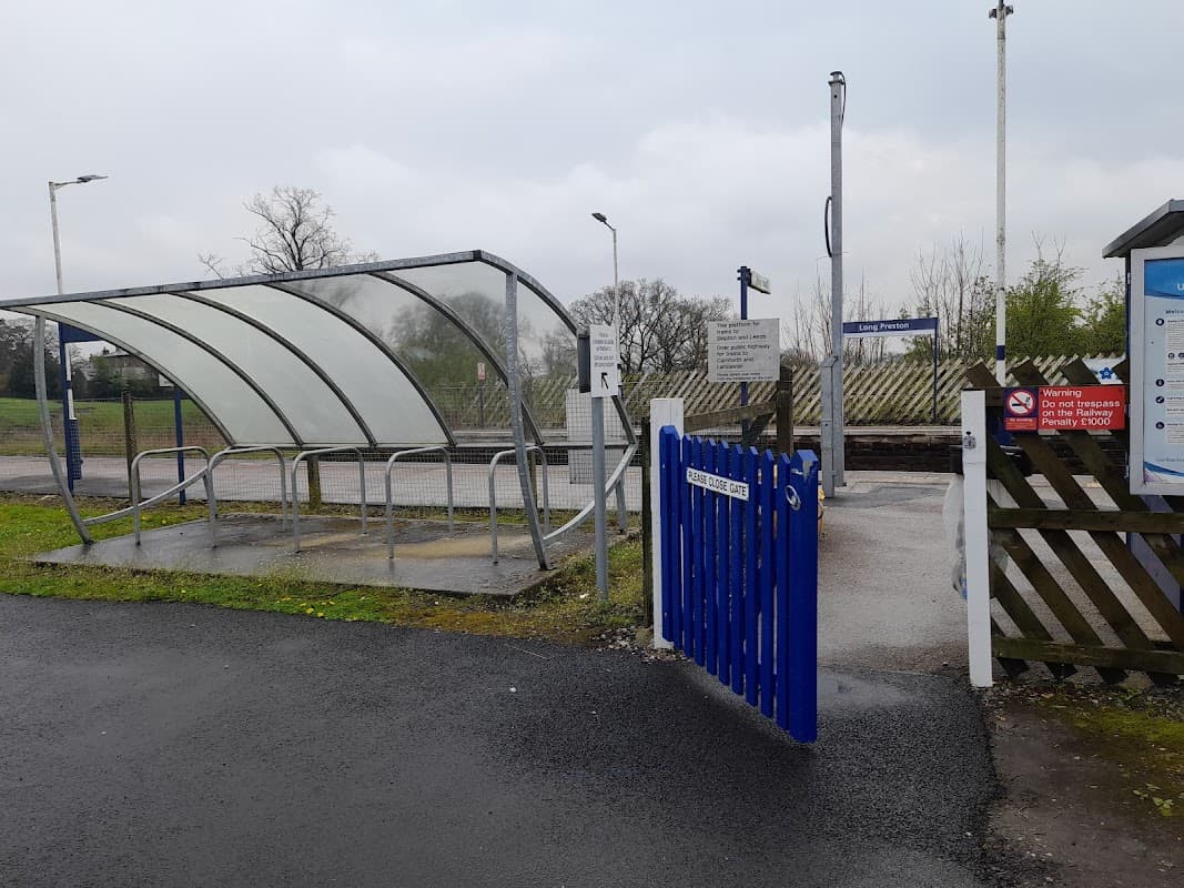 Bus stop shelter with a blue gate, signposts, and a grassy area in Long Preston, Yorkshire, under a cloudy sky.