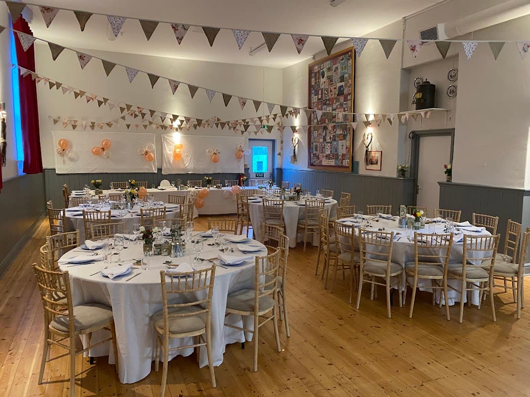 Decorated event space with round tables, chairs, bunting, and floral centerpieces, set for a celebration.