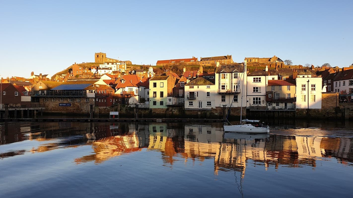 Colorful houses line the waterfront, reflecting in calm waters, with a hilltop church tower in the background.
