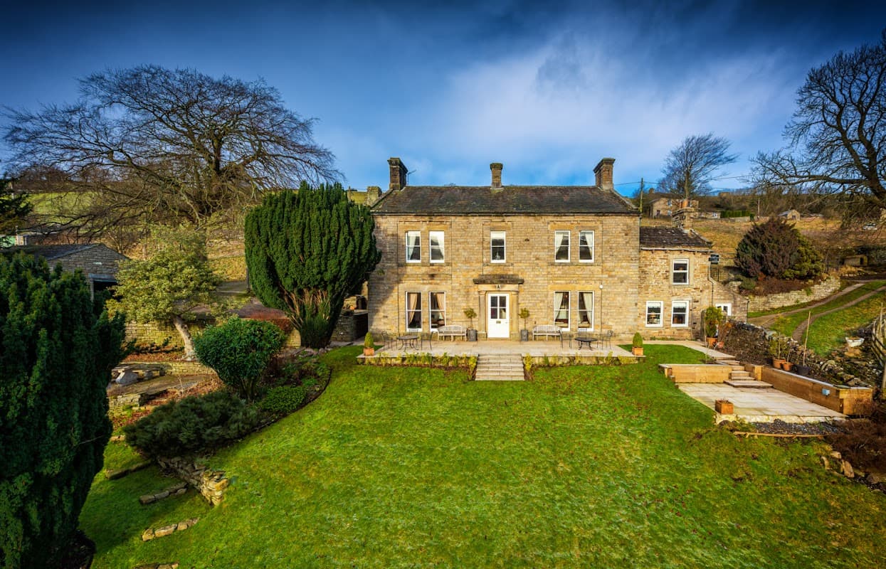 Stone building with large windows, surrounded by greenery and a well-maintained garden in a rural setting.