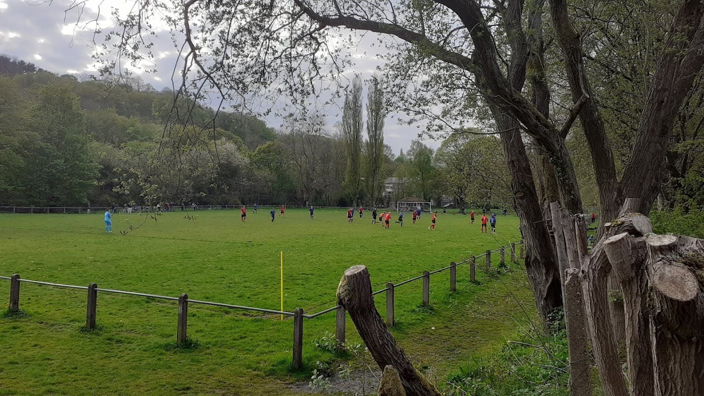A green football field with players in red jerseys, surrounded by trees and a wooden fence in Luddenden, Yorkshire.