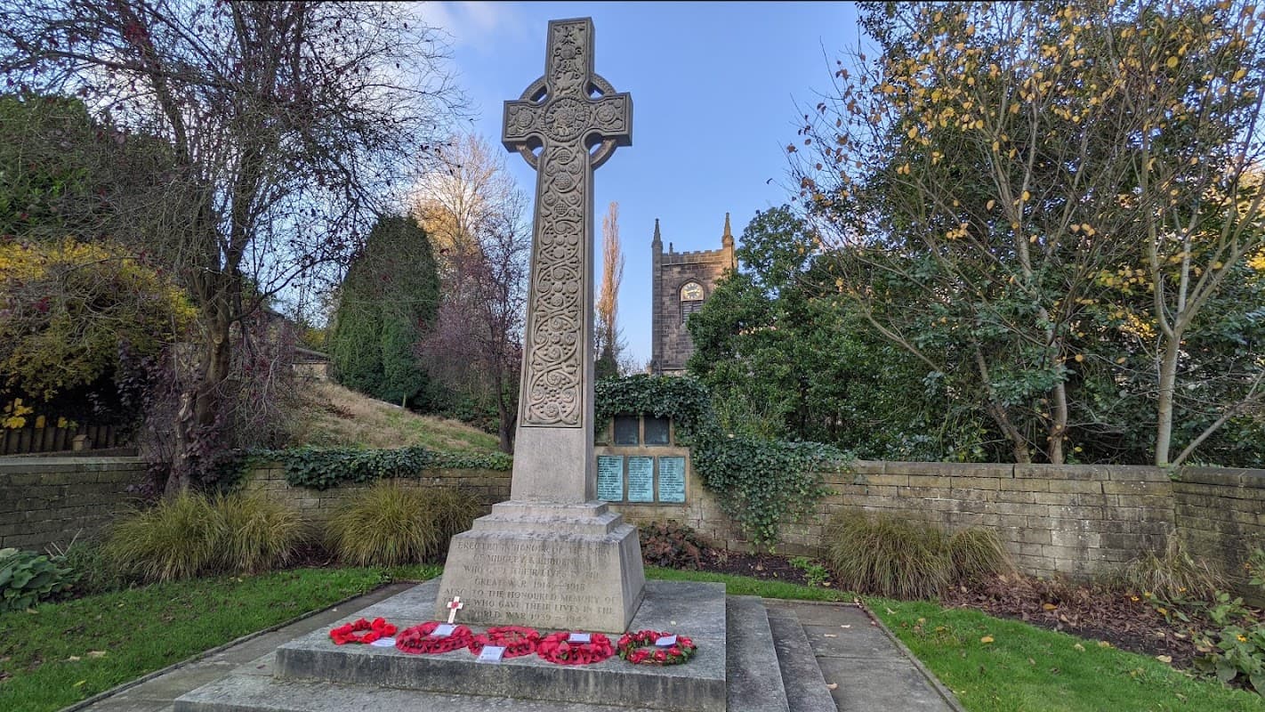 Celtic cross cenotaph with red poppy wreaths, surrounded by greenery and a stone wall, with a church in the background.
