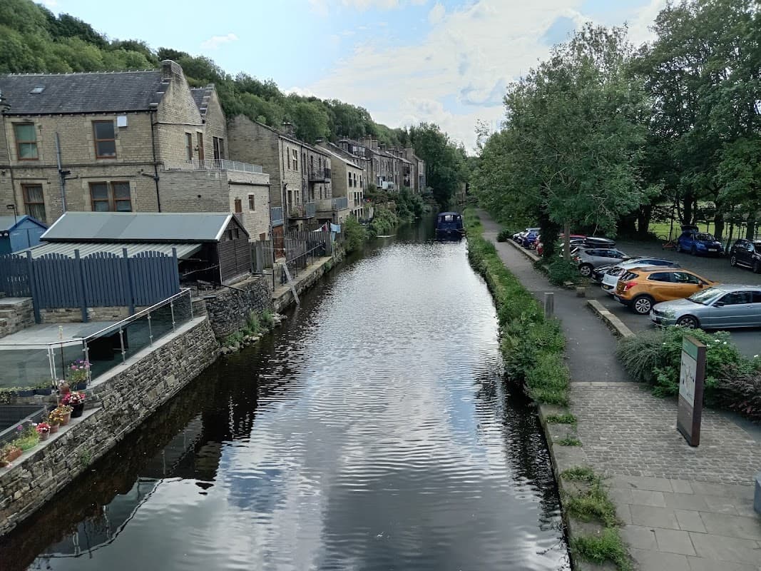 View of a canal beside Station Road car park, with stone buildings, greenery, and parked cars along the pathway.