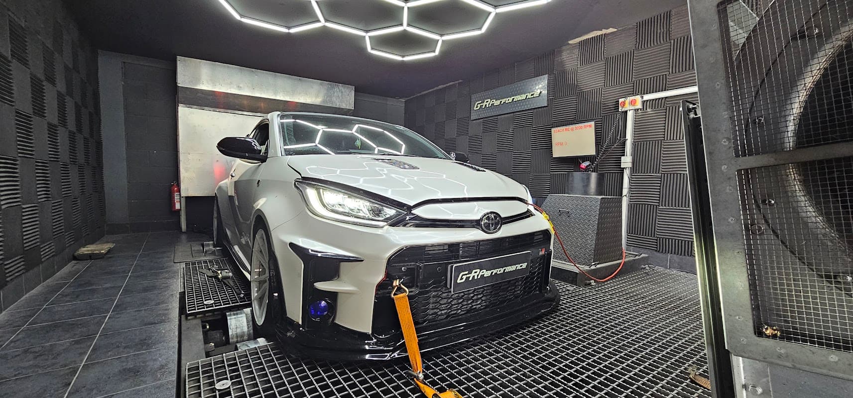 White Toyota car on a dynamometer in a high-tech performance testing facility with hexagonal ceiling lights.