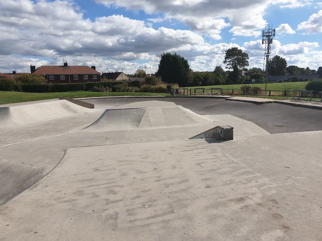 Concrete skatepark features ramps and bowls under a partly cloudy sky, surrounded by grass and houses in Maltby.
