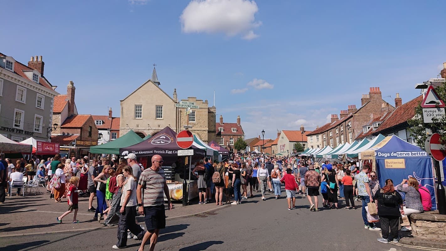 Busy market day in Malton, Yorkshire, with stalls, crowds, and historic buildings under a clear blue sky.