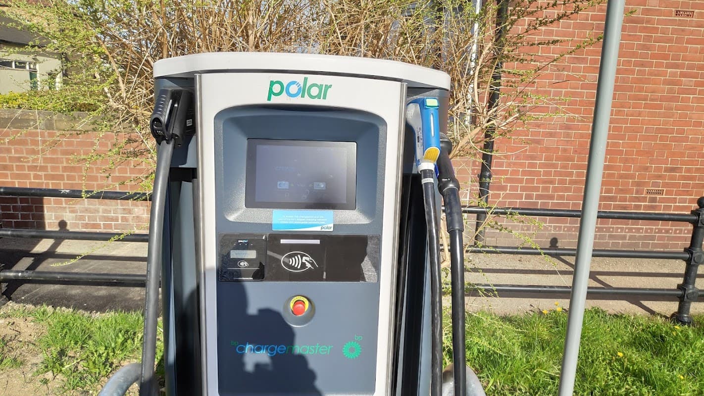 Electric vehicle charging station with a touchscreen and charging cables, set against a brick wall and greenery.
