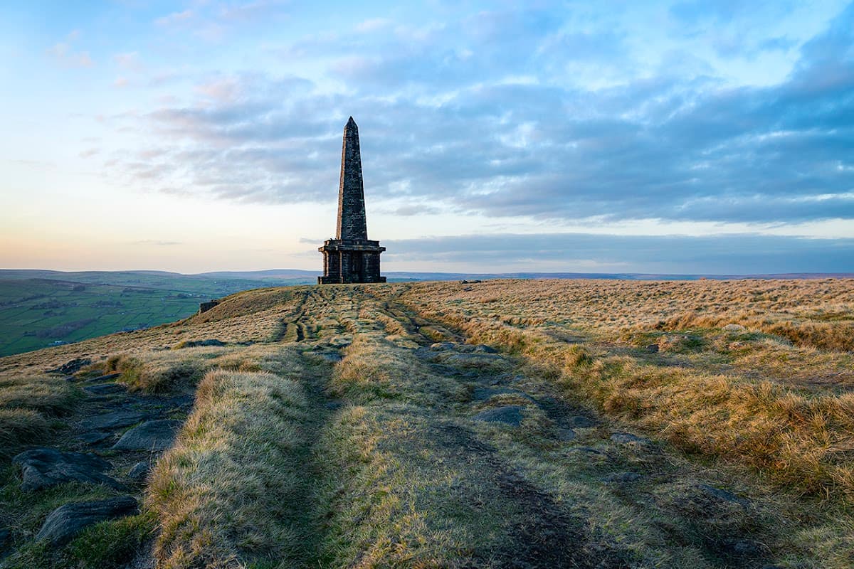 Tall stone monument atop grassy hill, surrounded by rolling hills and a cloudy sky at sunset.