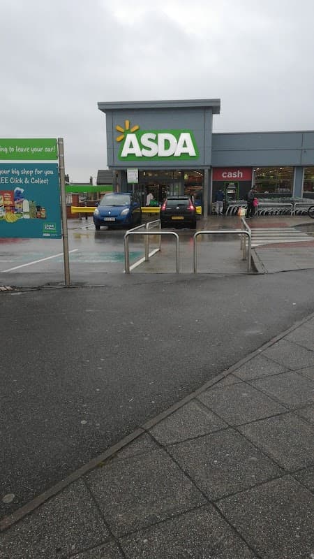 Asda supermarket entrance with green signage, cars parked in front, and shopping trolleys nearby on a cloudy day.