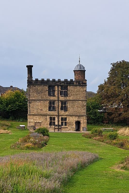 Historic stone building with a tower, surrounded by lush green lawn and flowering plants in Manor Lane Open Space.