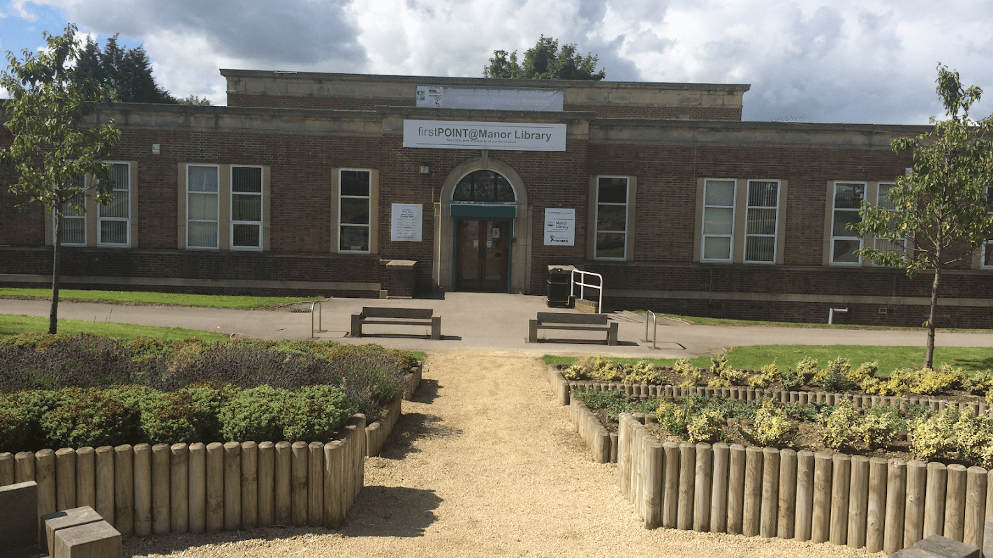 Brick building with large windows, green entrance, and landscaped garden featuring wooden borders and gravel paths.