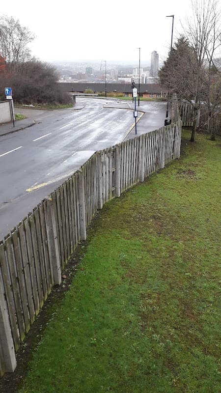 A quiet street with a wooden fence, grass, and a view of distant buildings under a cloudy sky.