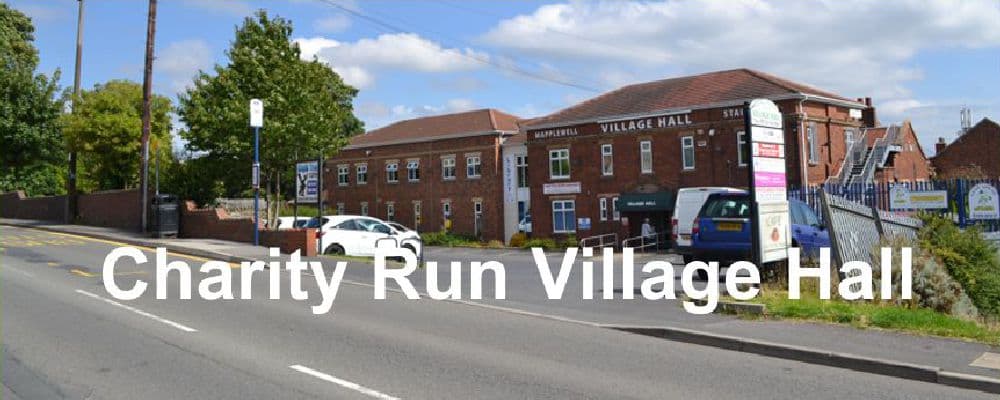 Village hall with red brick facade, sign reading "Charity Run Village Hall," parked cars, and green trees nearby.