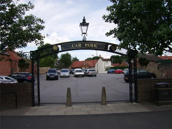 Entrance to Market Hill Car Park with a decorative arch, trees, and parked cars in a spacious lot.