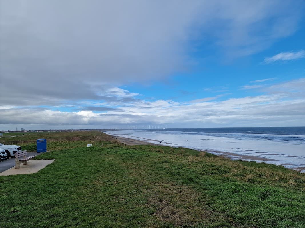 Coastal view from a grassy area with a car park, featuring a cloudy sky and distant shoreline along Marske.