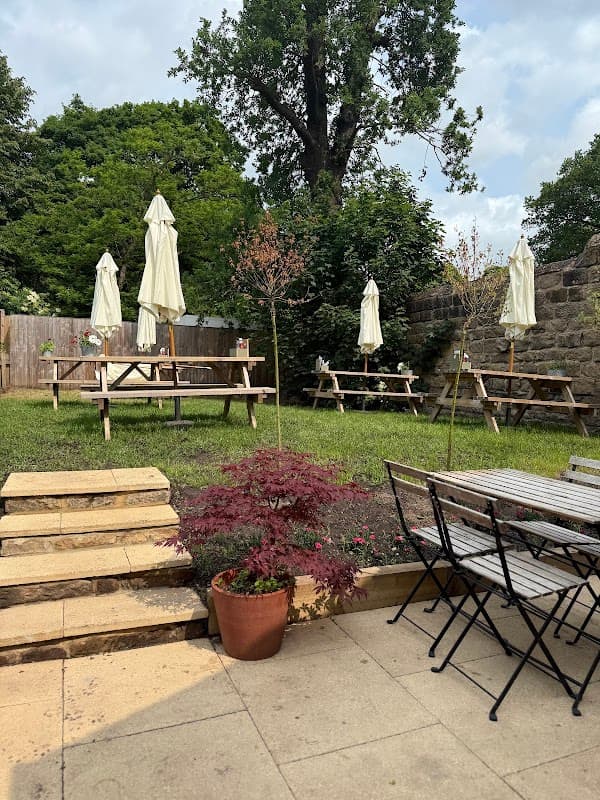 Three picnic tables with umbrellas, surrounded by greenery and a stone wall, with a potted plant in the foreground.