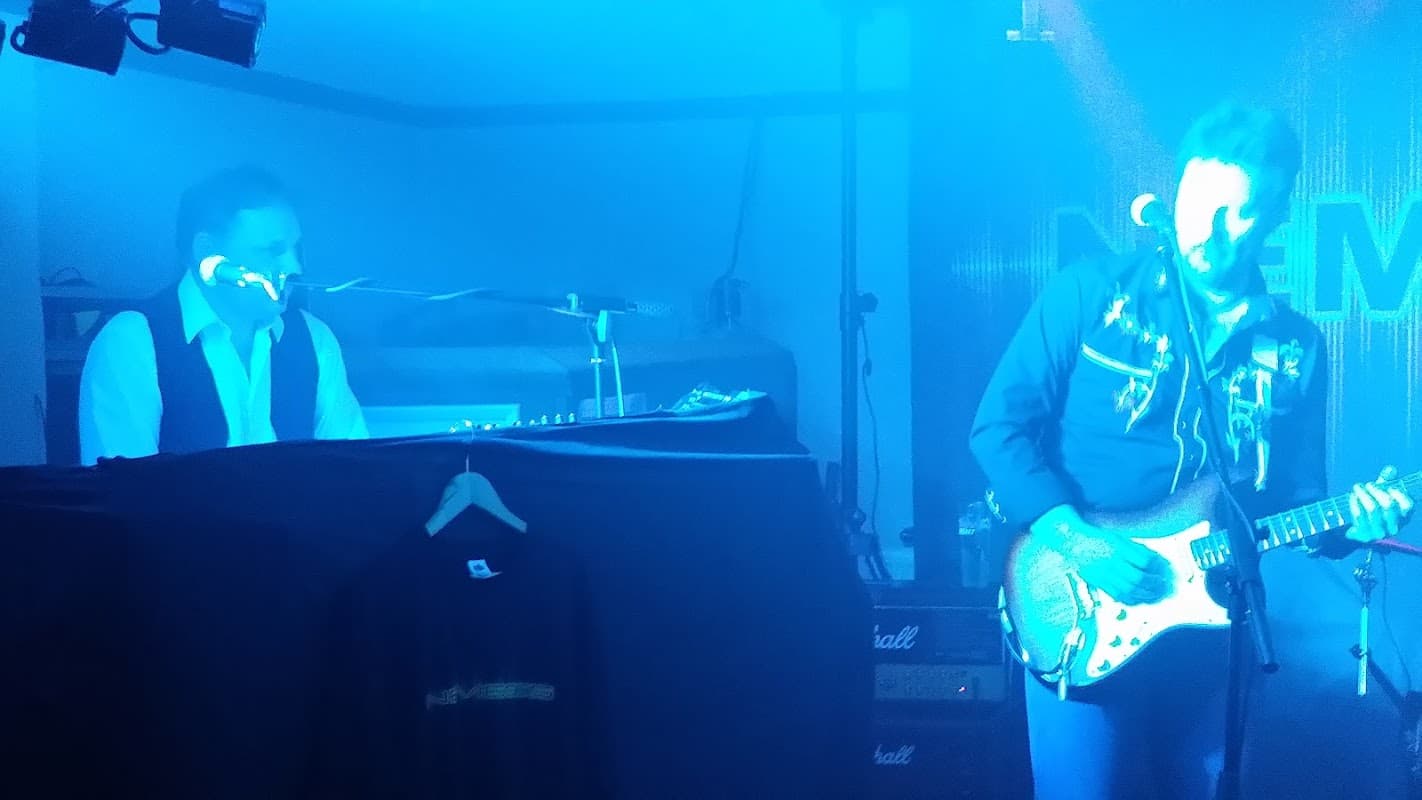 A band performing on stage with blue lighting; a keyboardist and a guitarist in focus.