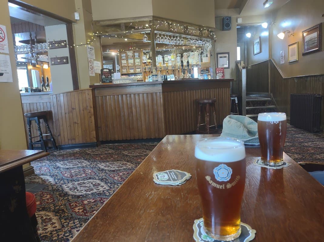 Two pints of dark beer on a wooden table, with a rustic bar and decorative lights in the background.