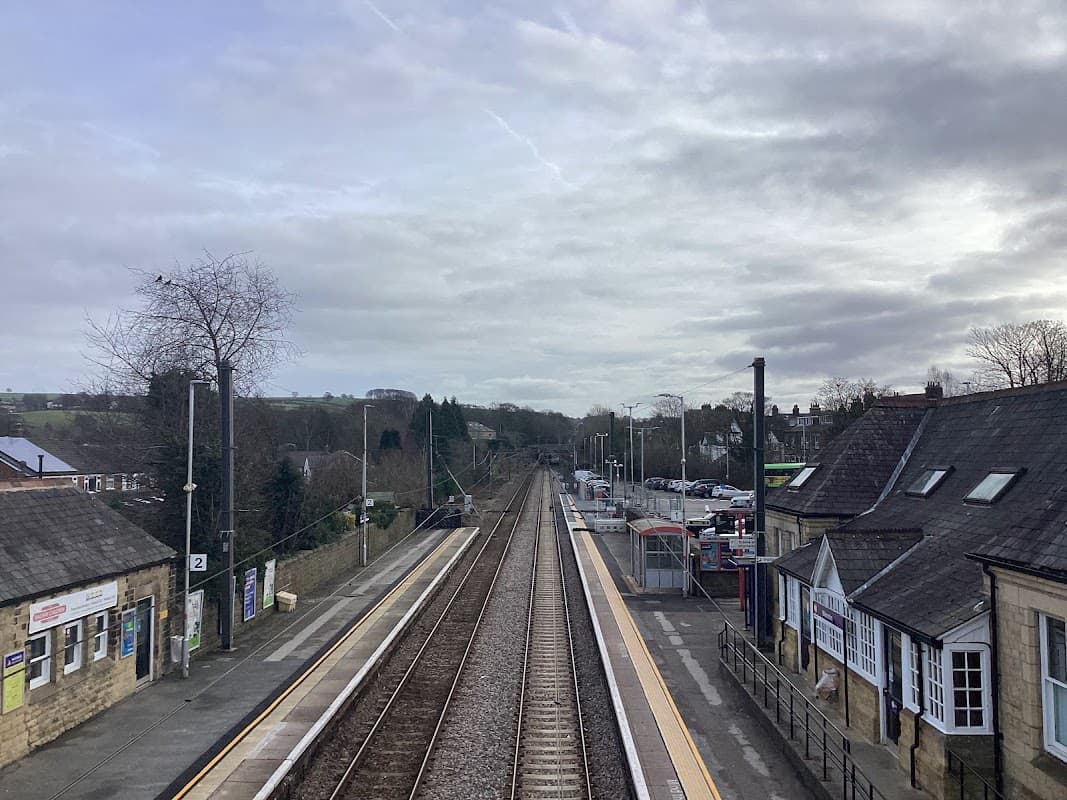 Train tracks stretch through Menston station, with buildings and trees lining the sides under a cloudy sky.