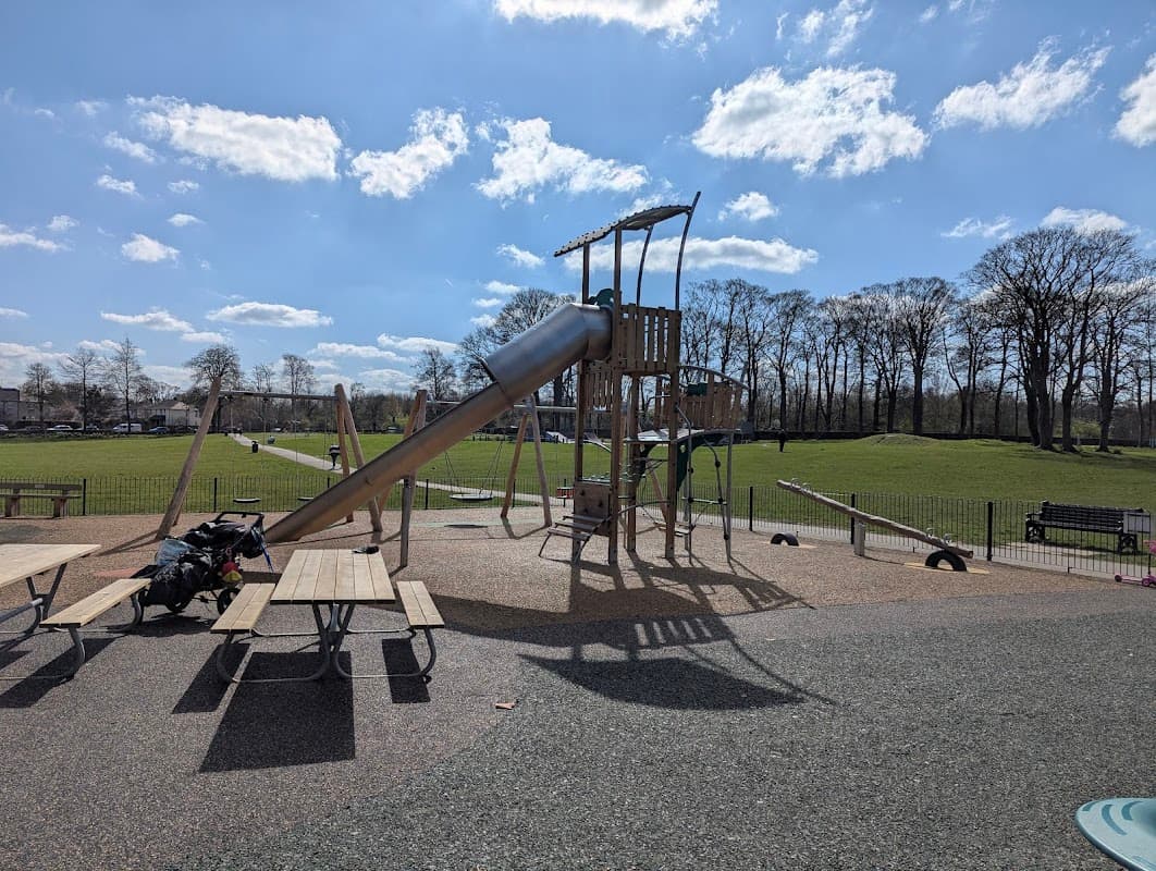 Playground with a slide, picnic tables, and green hills under a blue sky with fluffy clouds in Menston Park.