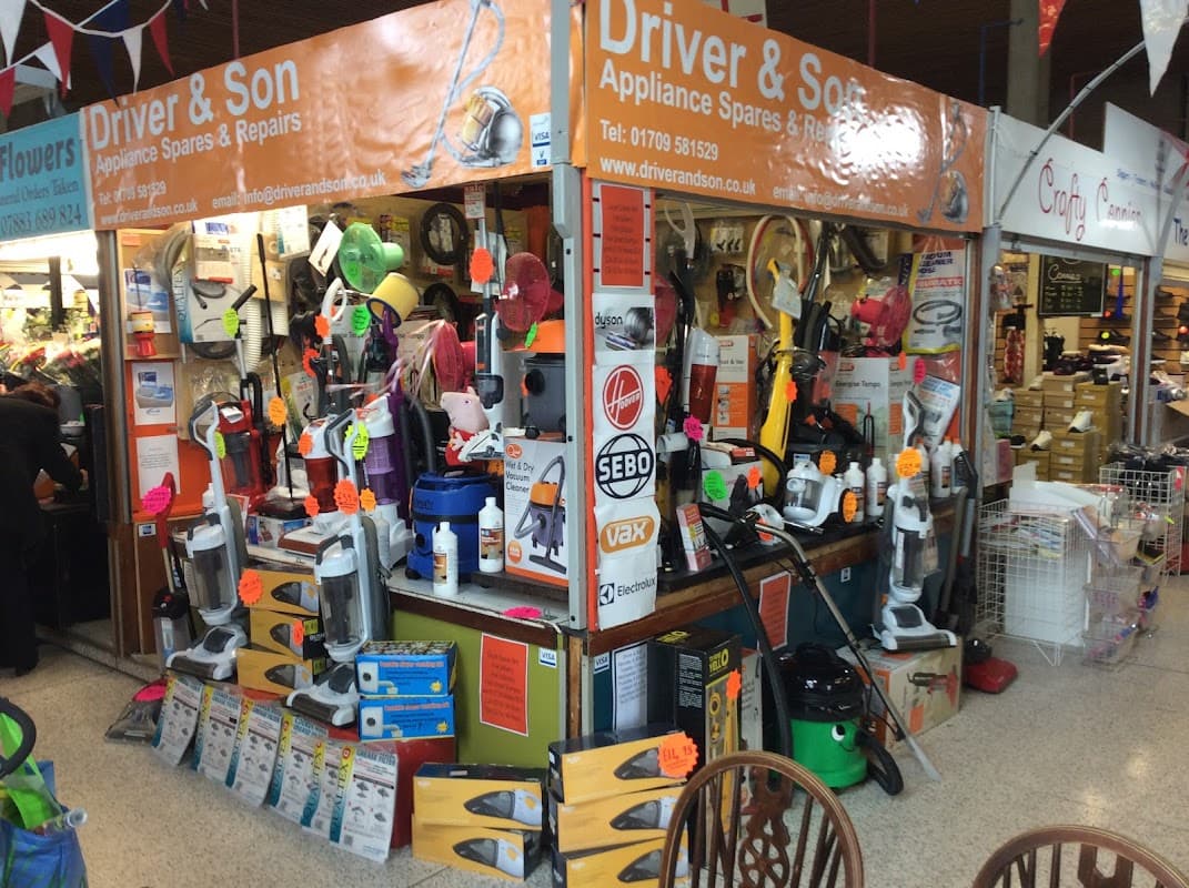 Market stall displaying various vacuum cleaners, appliance spares, and tools, with colorful signage and price tags.