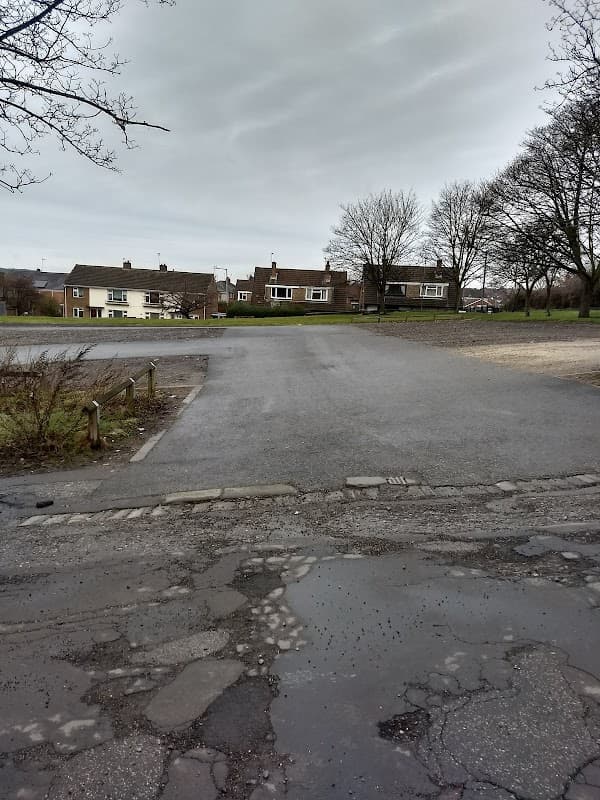 A gravel car park with a few trees, houses in the background, and a cloudy sky overhead.