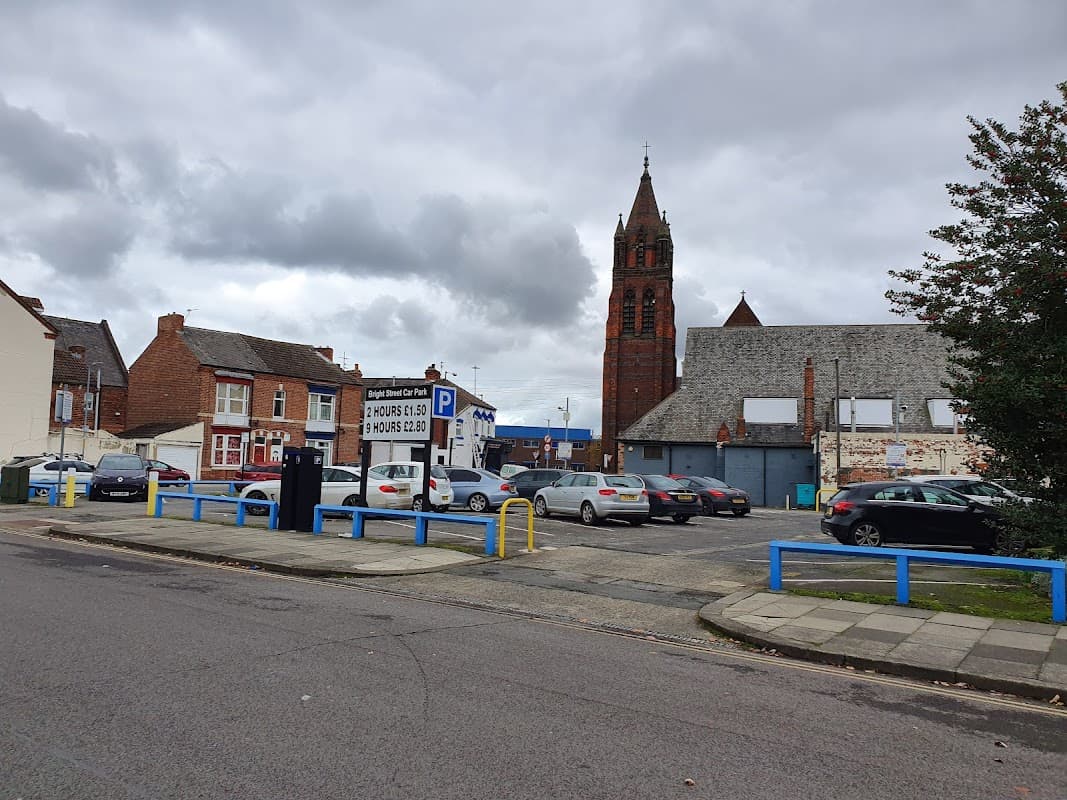 Pay & Display car park with parked cars, blue barriers, and a tall church tower in the background under a cloudy sky.