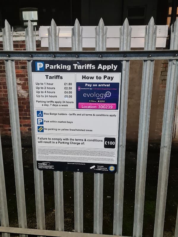 Parking tariff sign with payment instructions and rates displayed on a metal fence at a car park in Middlesbrough.