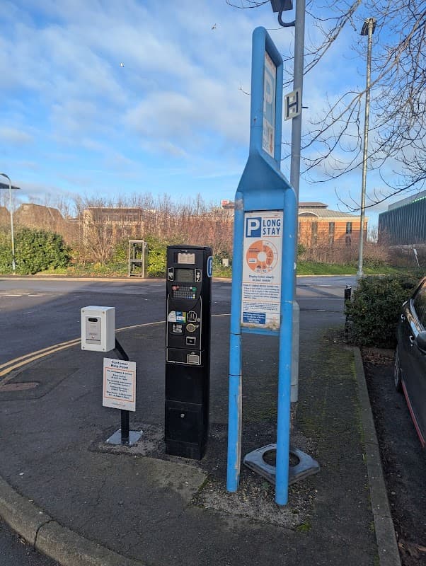 Pay & Display parking machine with signage, surrounded by greenery and buildings under a clear blue sky.