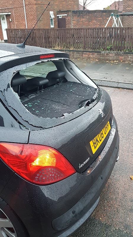 A black car with a shattered rear window parked on a wet street, with scattered glass and a residential fence in the background.