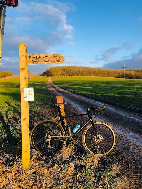 A bicycle leans against a wooden signpost on a dirt path surrounded by green fields and trees under a blue sky.