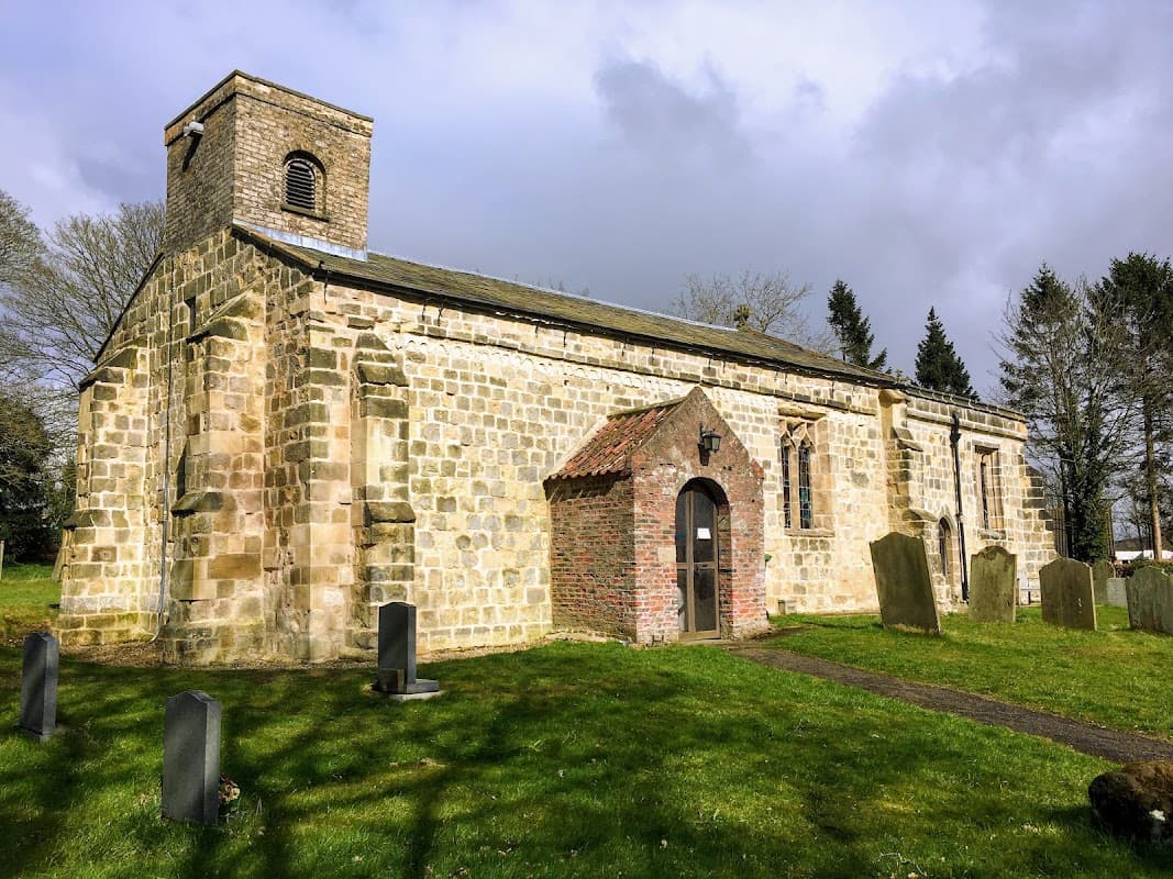Stone church with a bell tower, arched entrance, and surrounding graveyard in a green landscape under a cloudy sky.