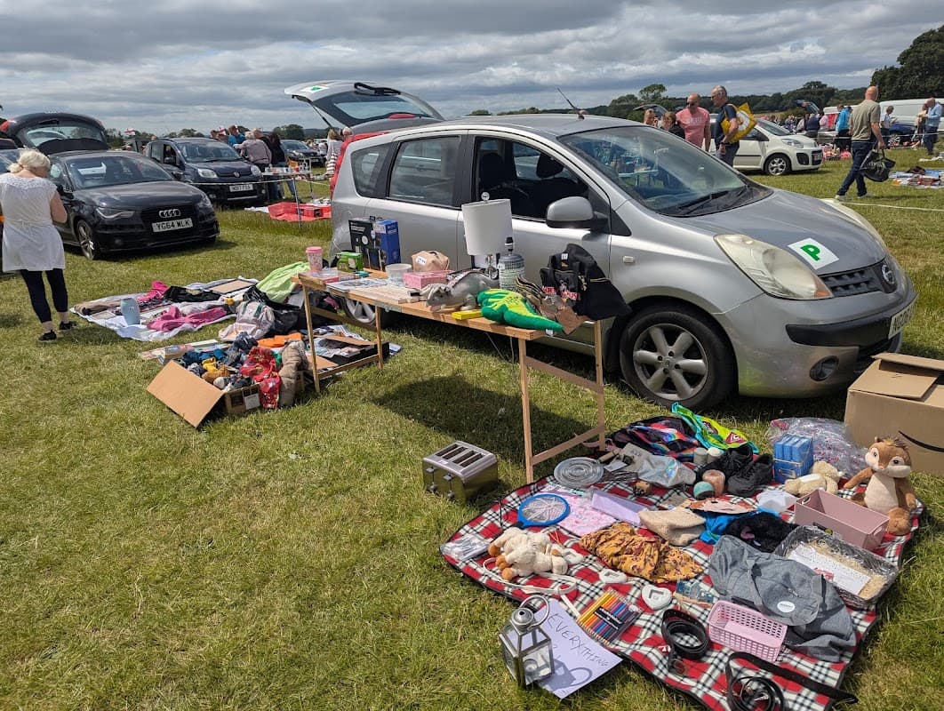 Car boot sale with various items displayed on tables, parked cars in the background, and people browsing in a grassy field.