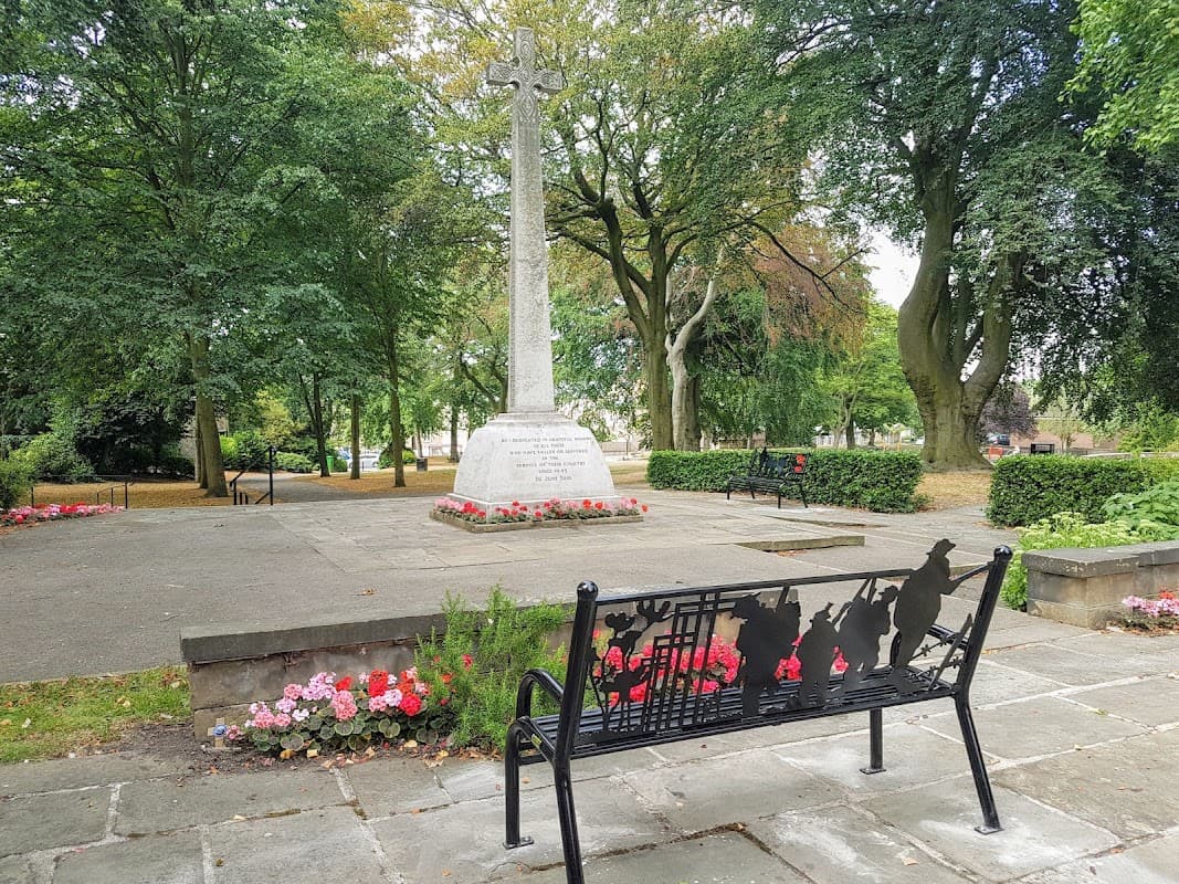 A memorial cross surrounded by trees and flower beds, with a decorative bench in the foreground.