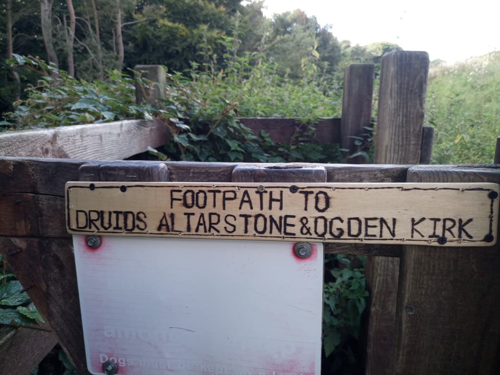 Signpost directing to footpath for Druids Altarstone and Ogden Kirk, surrounded by greenery and wooden fencing.