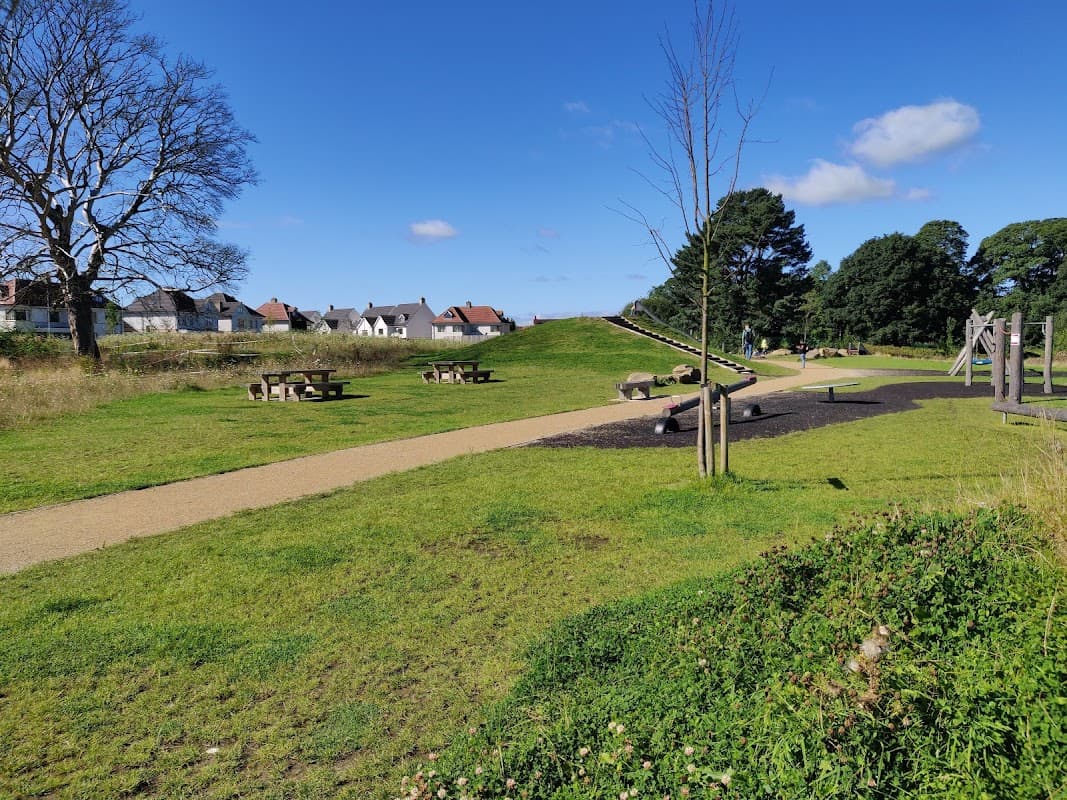 Lush green park with pathways, picnic tables, trees, and residential buildings under a clear blue sky.