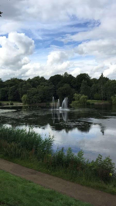 Tranquil lake with fountains, surrounded by lush greenery and cloudy blue sky in Roundhay Park, Moortown.