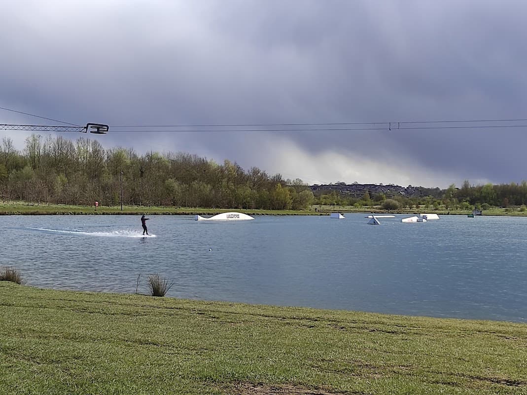 A person water skiing on a lake with a cable system, surrounded by greenery and dark clouds in the background.
