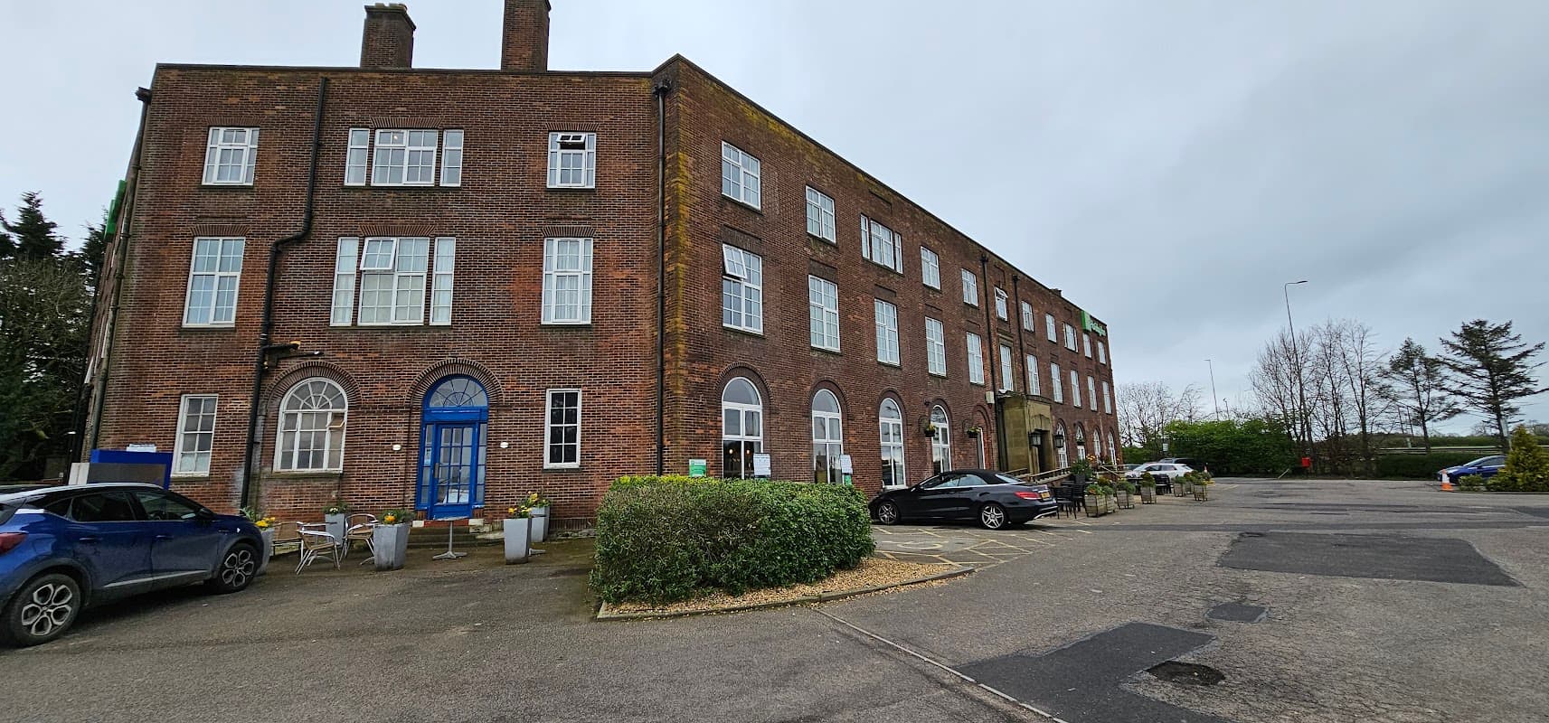 Tesla Supercharger station with a brick building, parked cars, and greenery in Moulton, North Yorkshire.