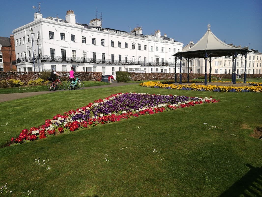 Colorful flower beds in geometric patterns, a gazebo, and people biking near a historic white building.