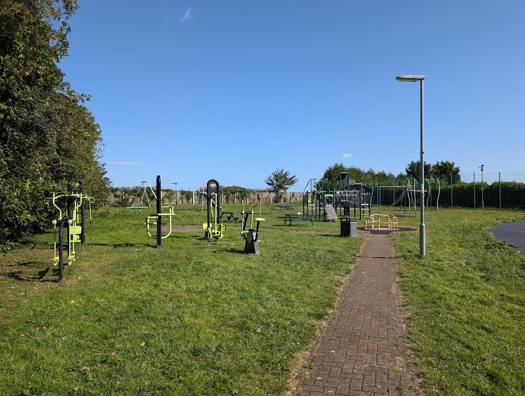 Outdoor play area with exercise equipment, playground structures, benches, and green grass under a clear blue sky.