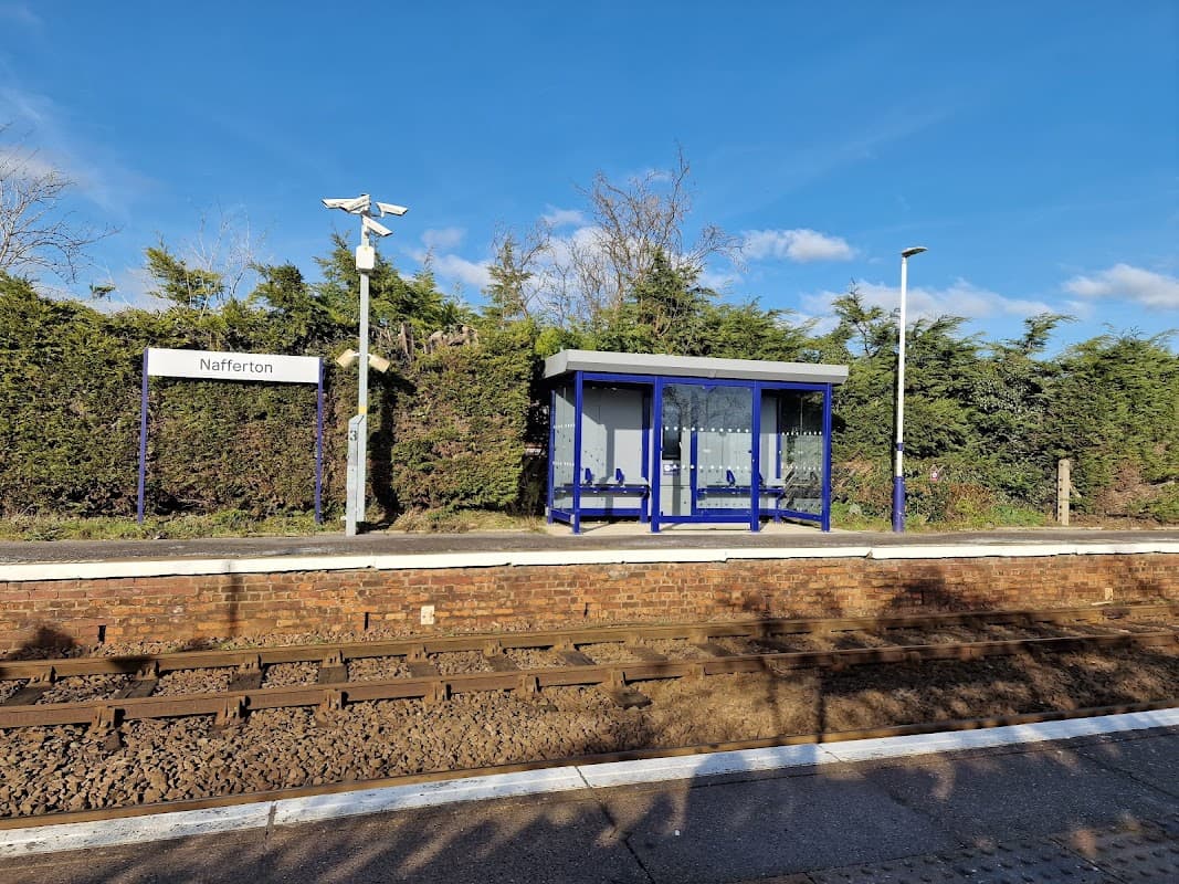 Nafferton train station with a blue shelter, sign, and overgrown greenery under a clear blue sky.