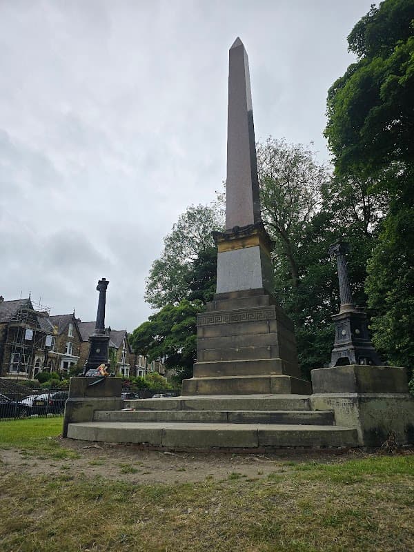 Tall stone obelisk surrounded by greenery, with ornate lampposts nearby and residential buildings in the background.