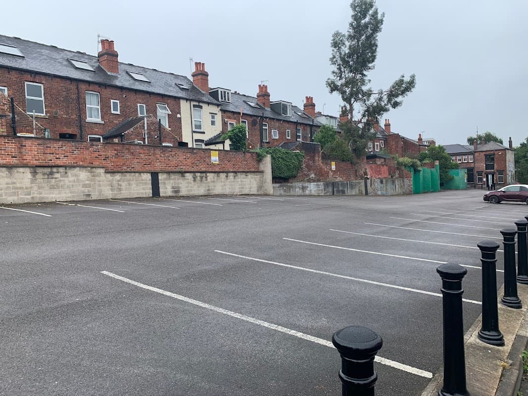Empty car park with brick buildings and chimneys in the background, surrounded by greenery and a cloudy sky.