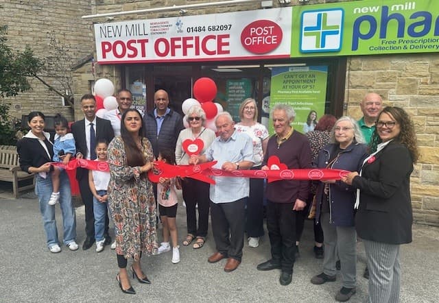 A ribbon-cutting ceremony at New Mill Post Office with a diverse group of people holding balloons and smiling.
