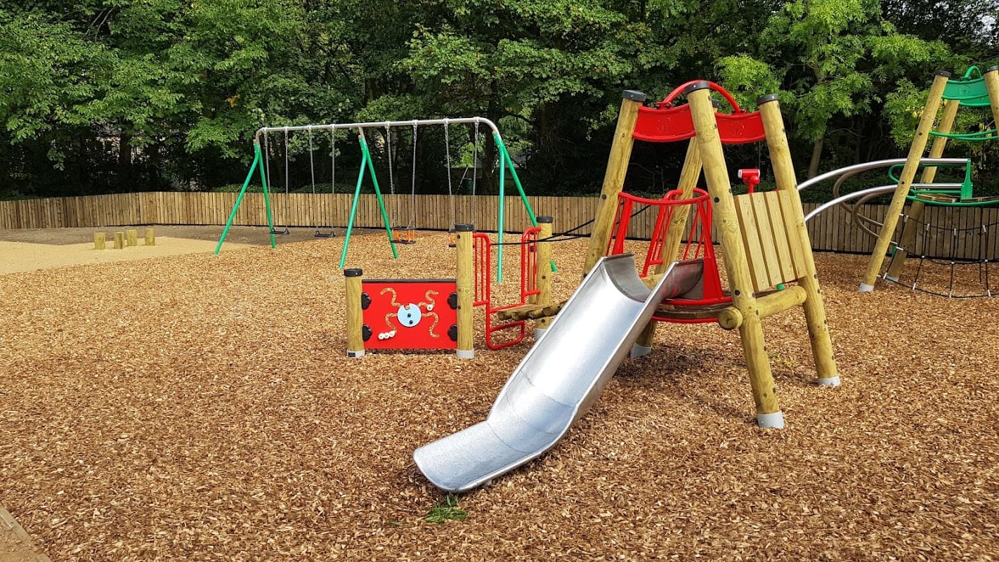 Playground featuring a silver slide, swings, climbing structures, and wood chip flooring surrounded by greenery.